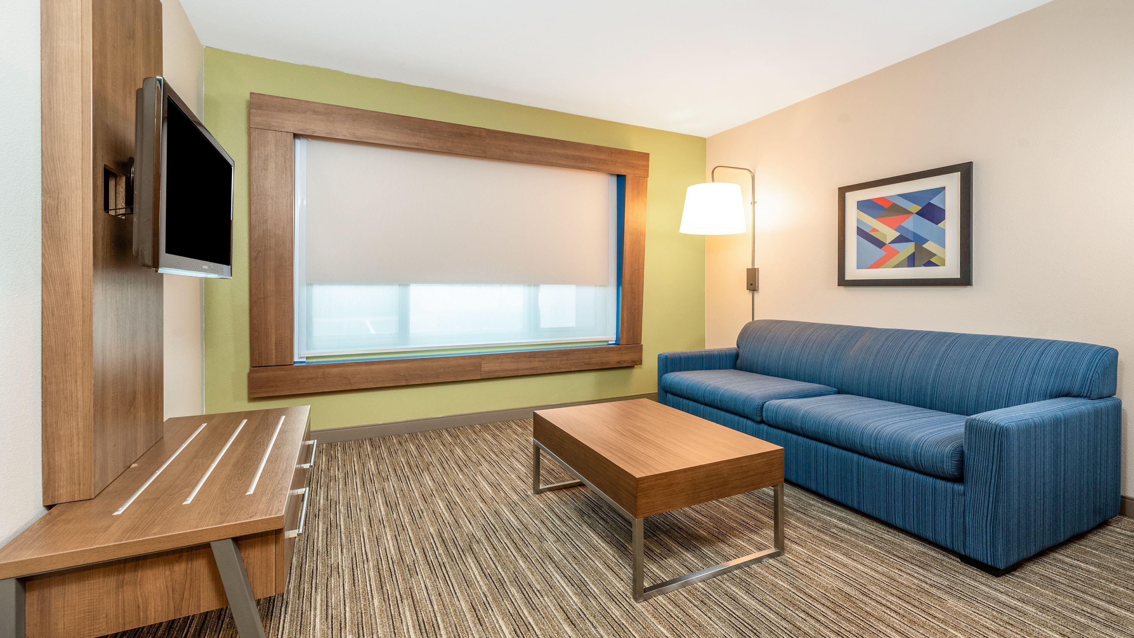 A hotel room with a wooden desk, a blue sofa, a TV mounted on a wooden panel, a large window with blinds, a floor lamp, and a framed abstract painting.