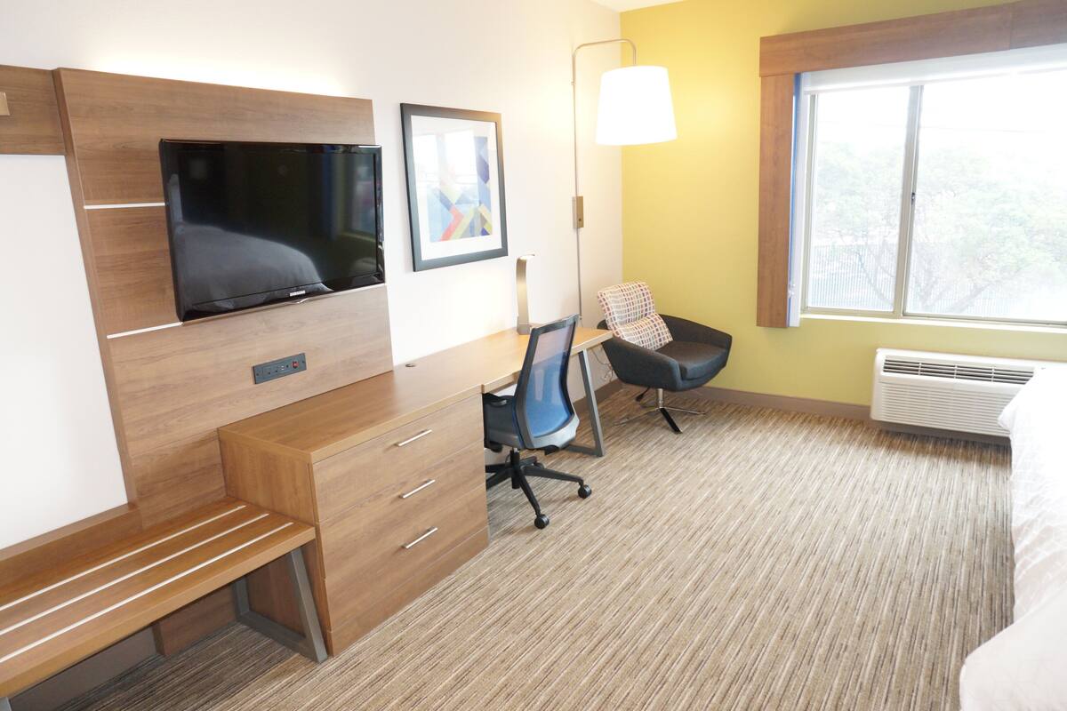 A hotel room with a wooden desk, a chair, a television, a lamp, and a window with a view.