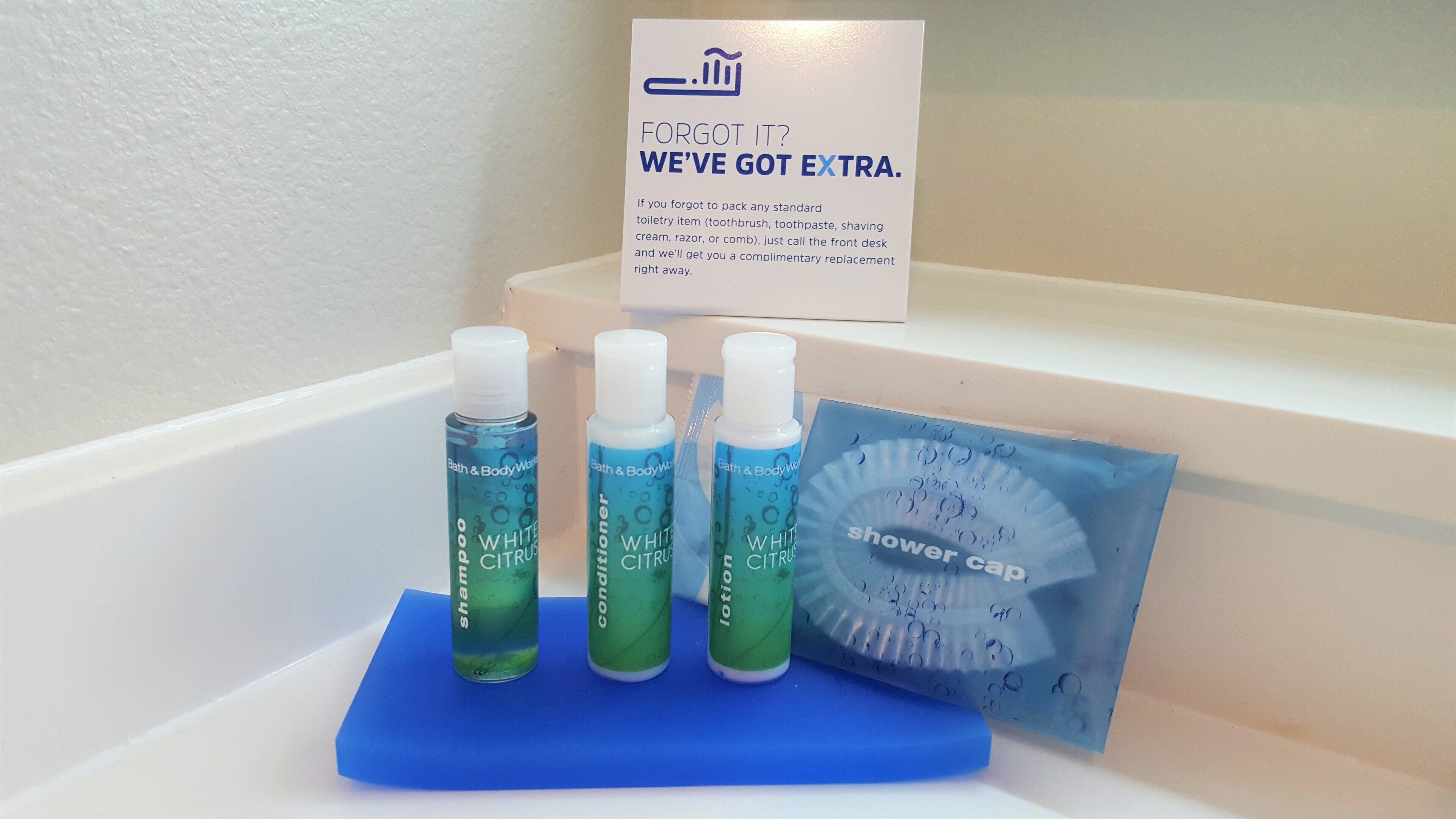 A bathroom counter with toiletries including shampoo, conditioner, lotion, and a shower cap. A sign above the counter reads "Forgot it? We've got extra."