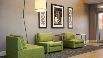 A hotel lobby with green armchairs, a floor lamp, framed artwork on the wall, and a small table.