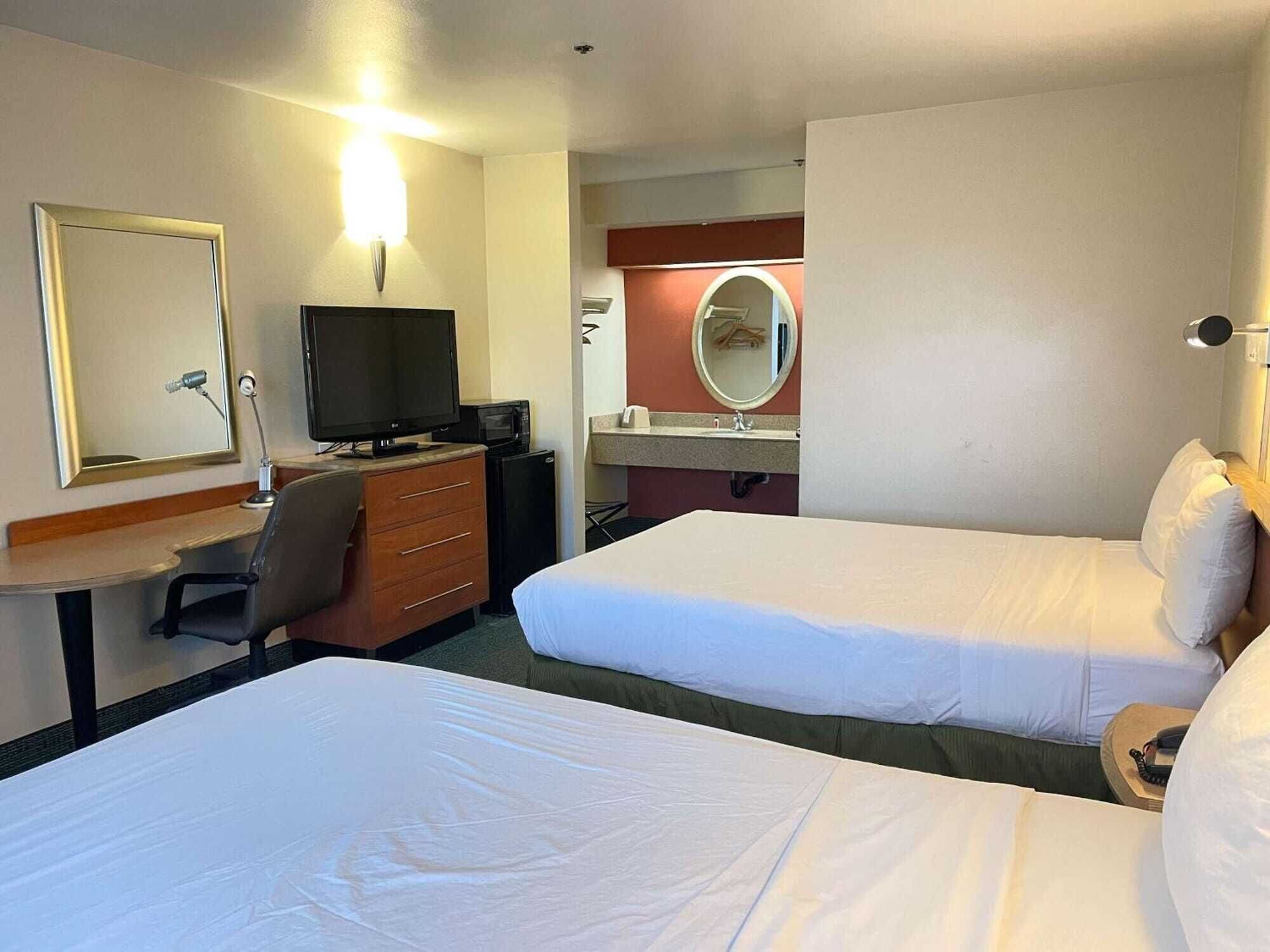 A hotel room with two beds, a desk with a computer, a television, a mirror, and a small kitchenette.