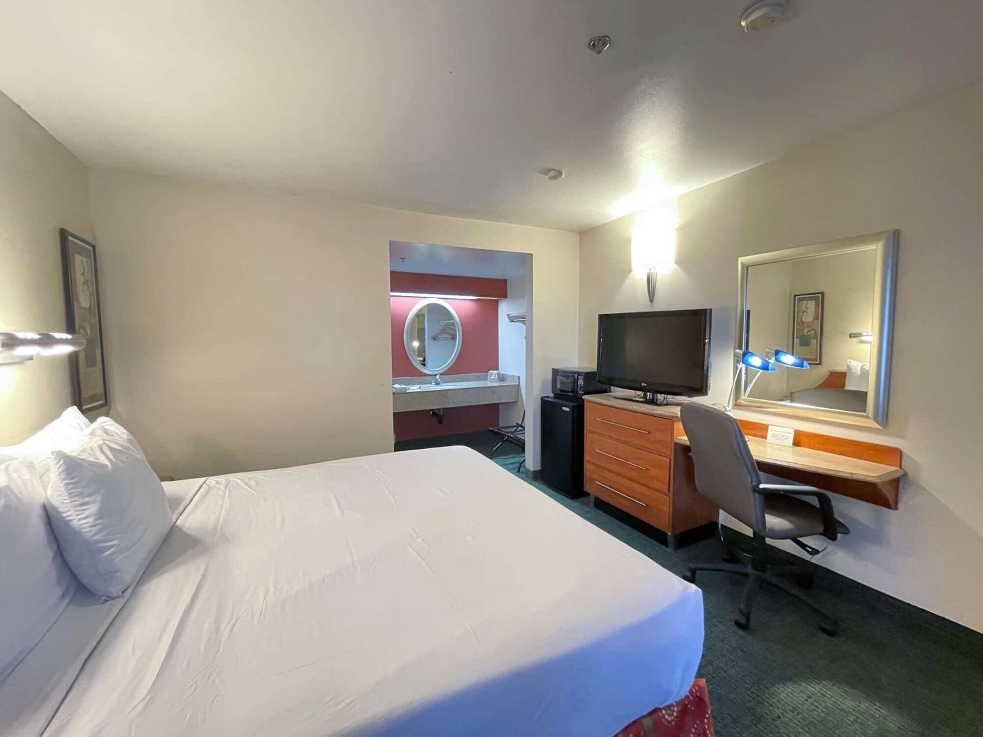 A hotel room with a bed, a desk with a computer, a television, a mirror, and a small kitchenette.