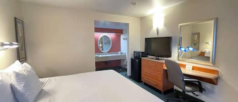 A hotel room with a bed, a desk with a computer, a TV, a mirror, and a small kitchenette.