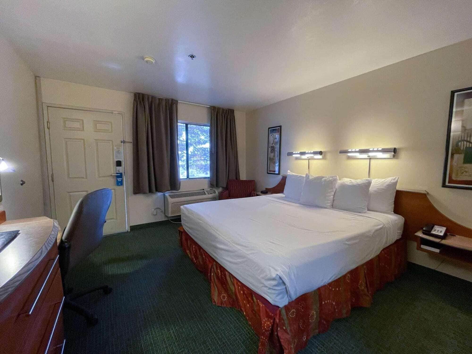A hotel room with a large bed, a desk, a chair, a window with curtains, and a wall-mounted lamp.