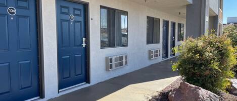 A row of blue doors with numbers 104 and 105, a small air conditioning unit, and a large rock in a landscaped area.
