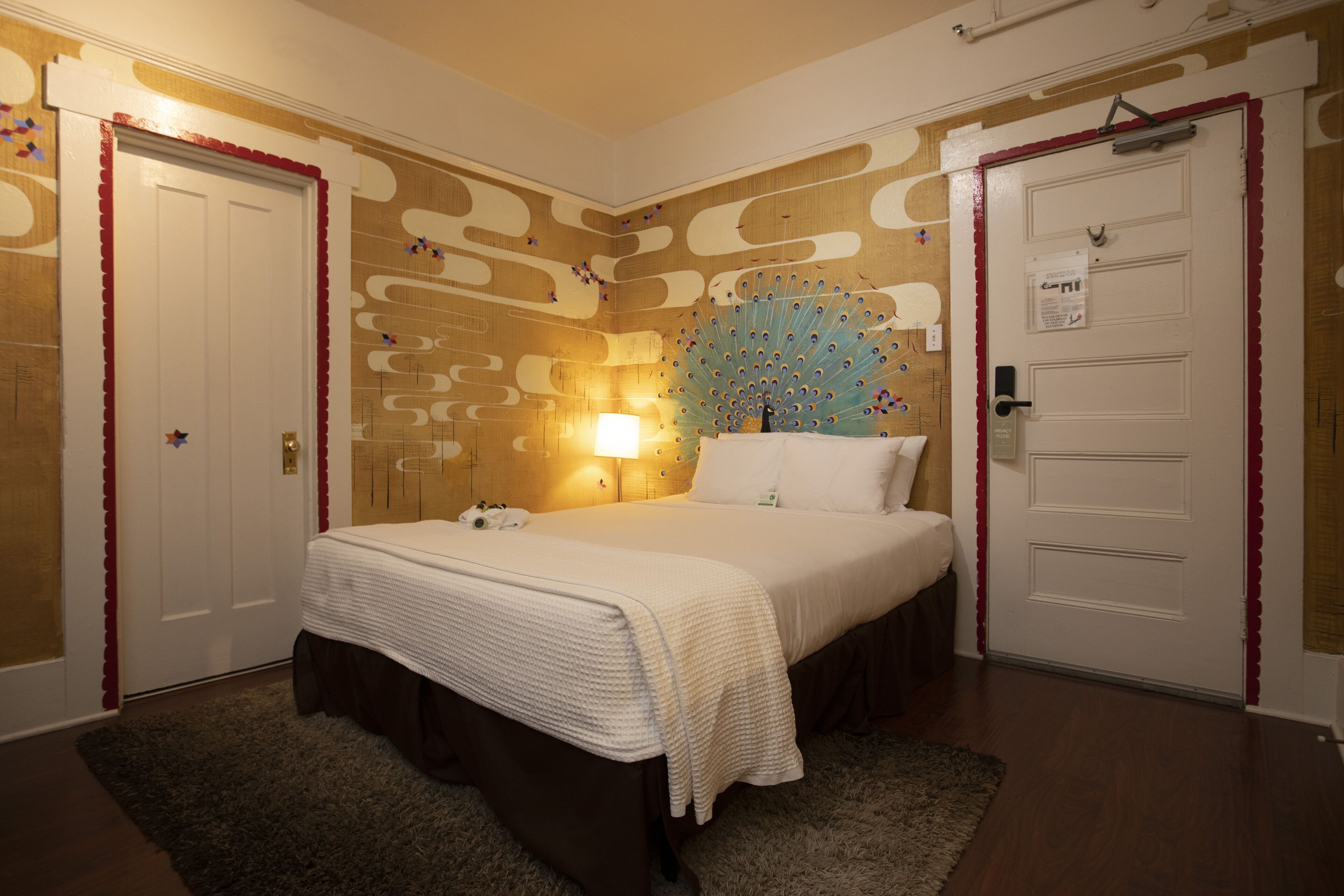 A hotel room with a bed, a nightstand, a wall clock, and a door with a red trim.