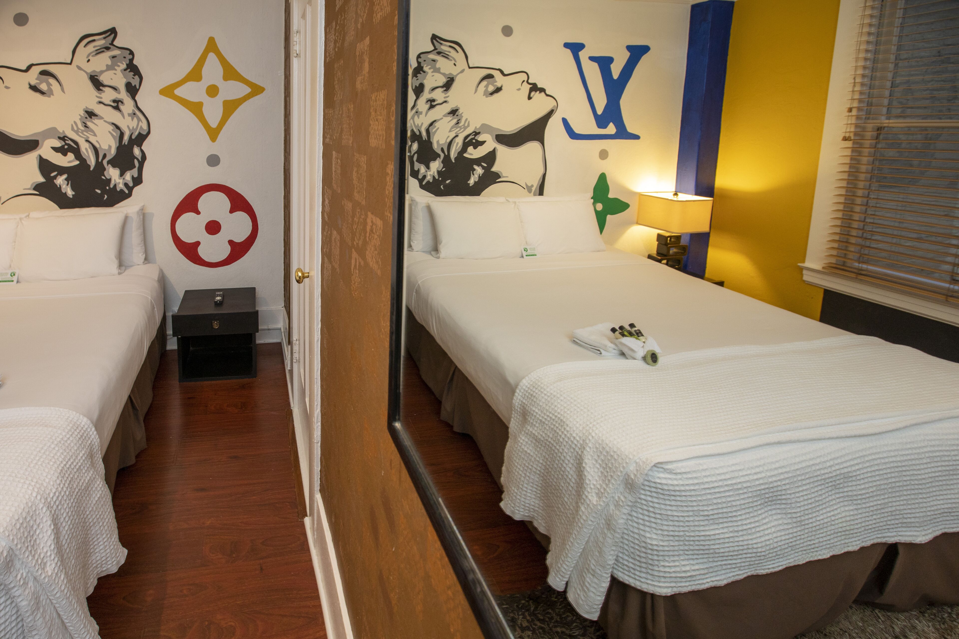 A hotel room with two single beds, a wooden floor, a mirror, and a wall with decorative elements including a dog, a flower, and a letter 'V'.