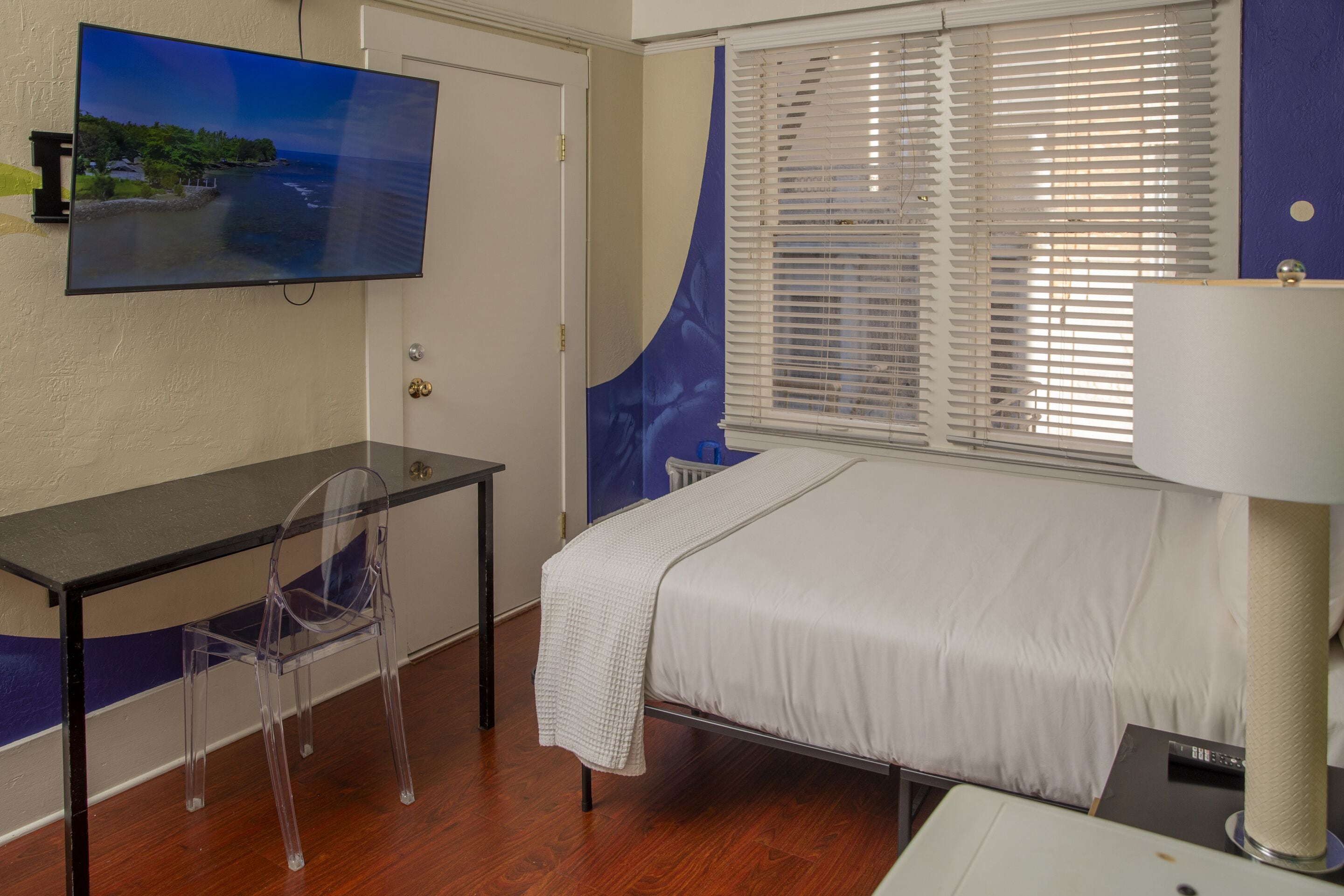 A hotel room with a bed, a desk with a chair, a TV mounted on the wall, and a window with blinds.