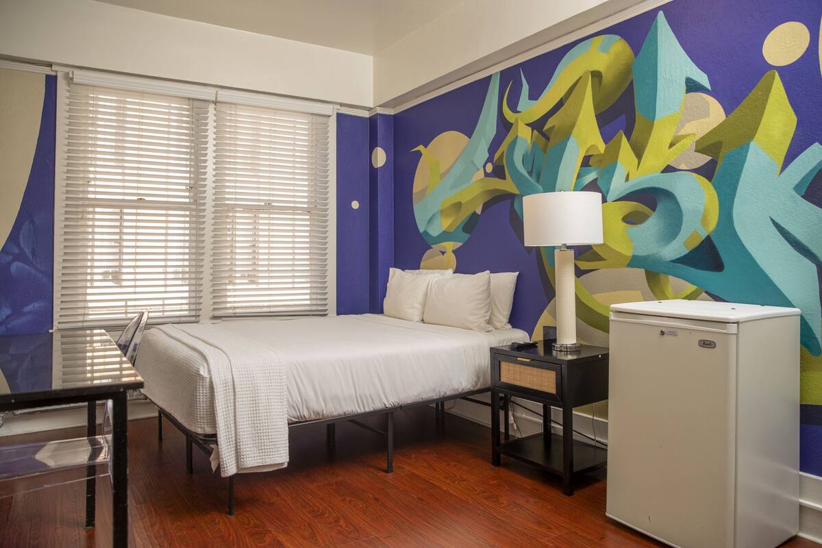 A hotel room with a bed, a desk, a lamp, and a refrigerator.