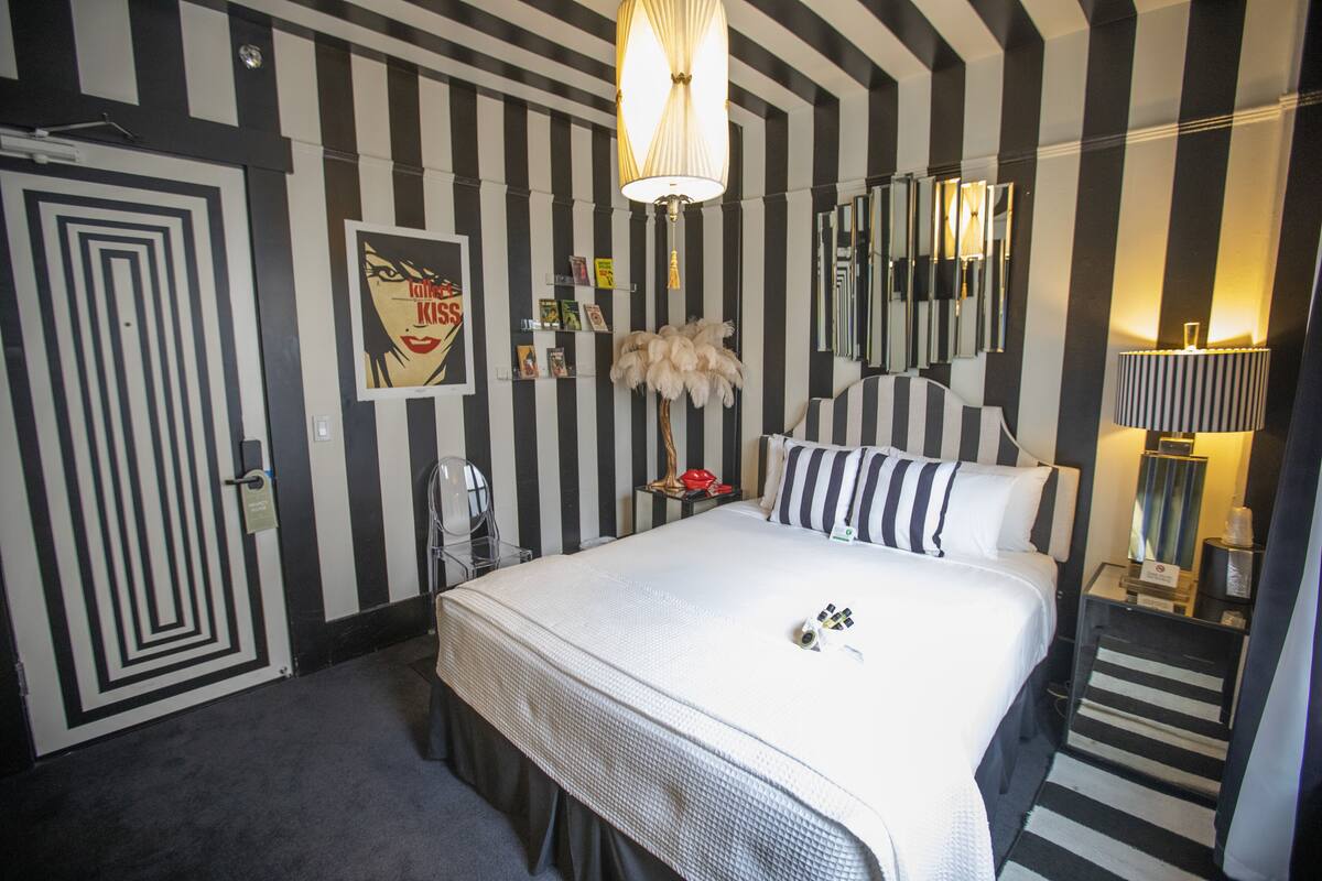A hotel room with a striped wall design, a large bed with white bedding, a mirror, a lamp, and a door with a unique pattern.