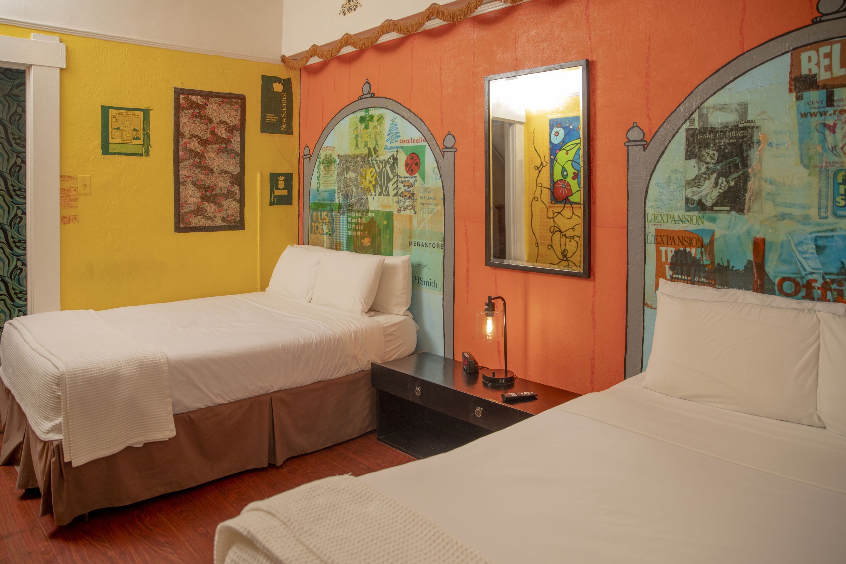 A room with two beds, a nightstand with a lamp, and a wall decorated with various posters and a mirror.
