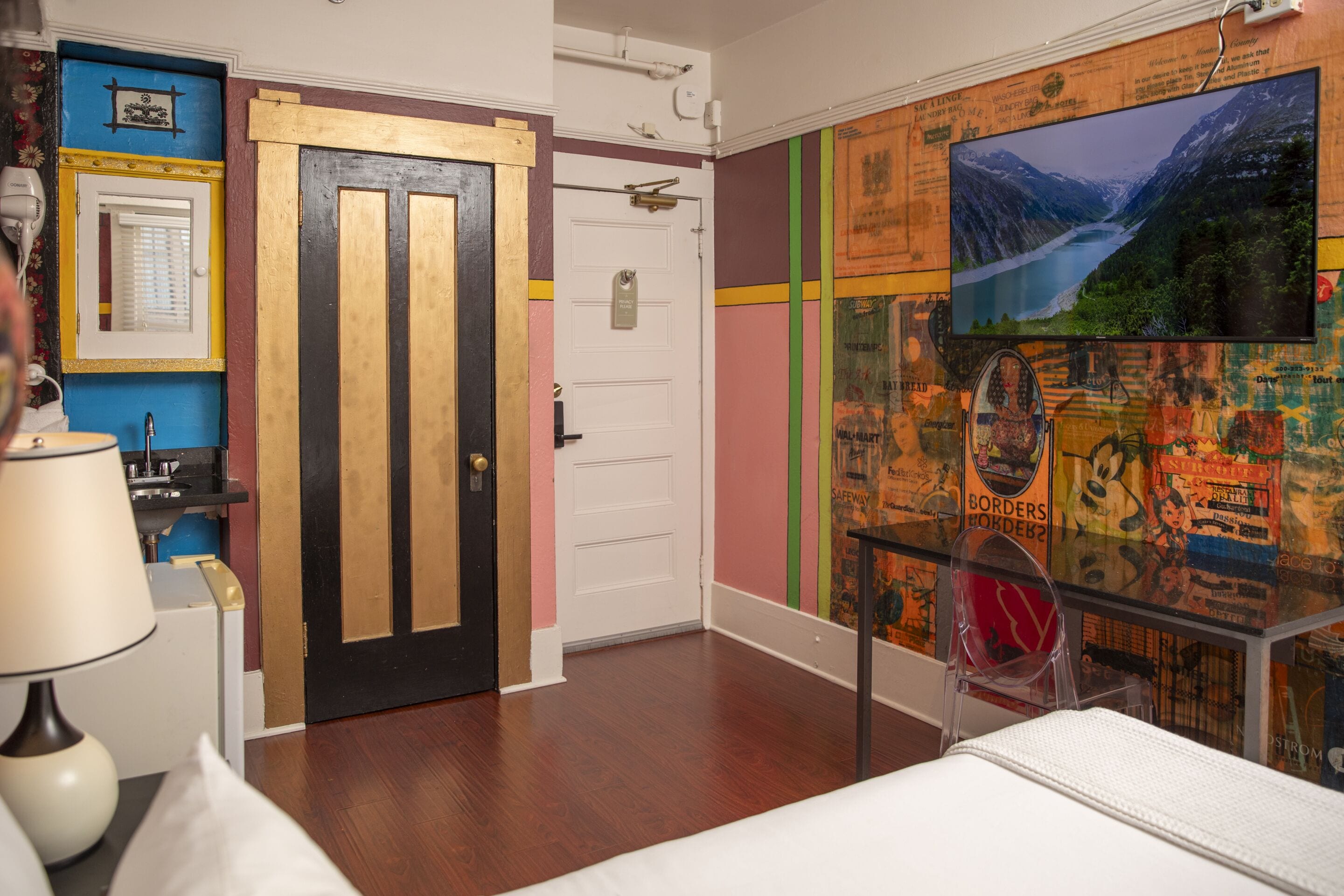 A room with a wooden door, a bed, a glass table, and a wall with various posters and a TV screen.