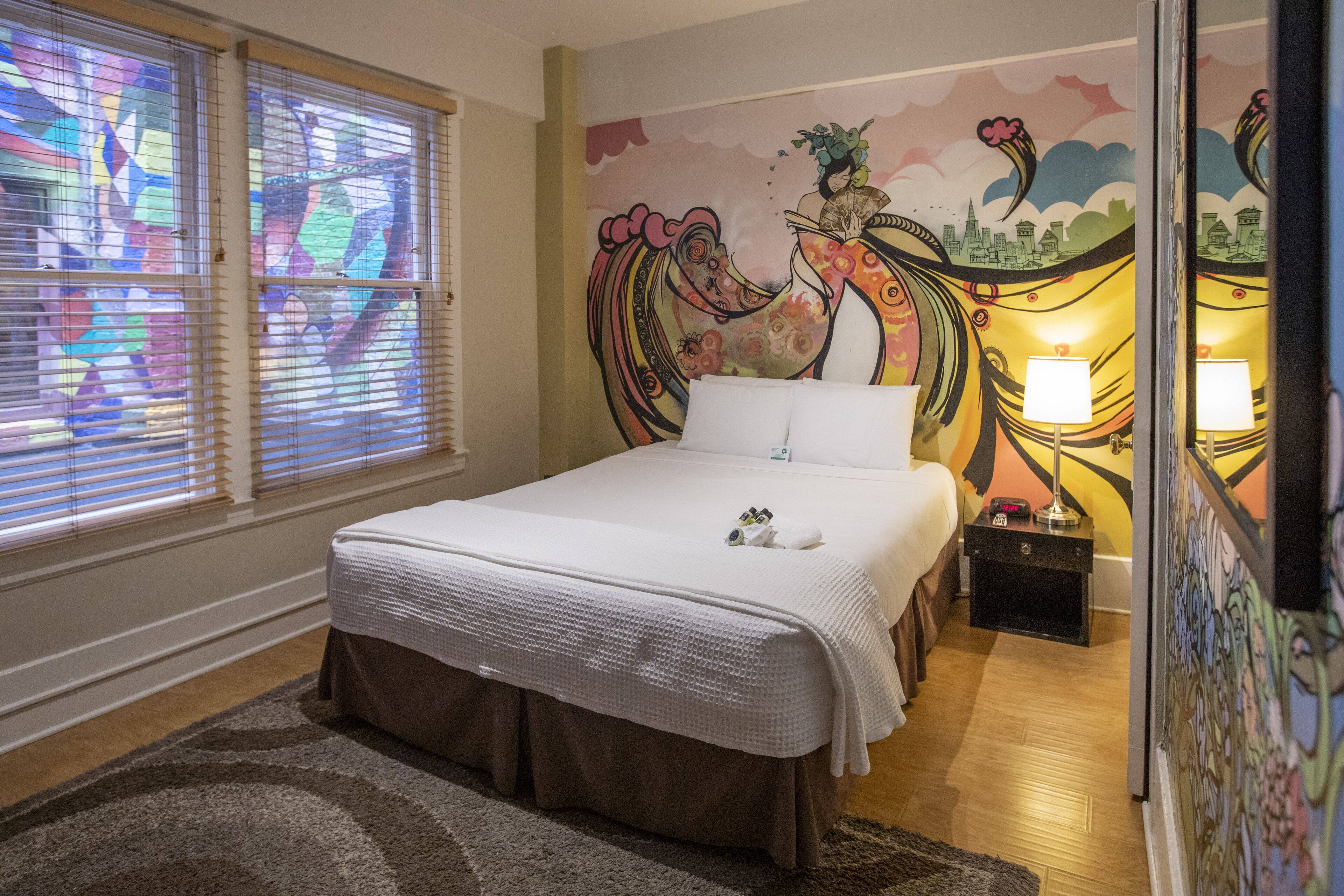 A bedroom with a bed, a nightstand, a lamp, and a colorful mural on the wall.