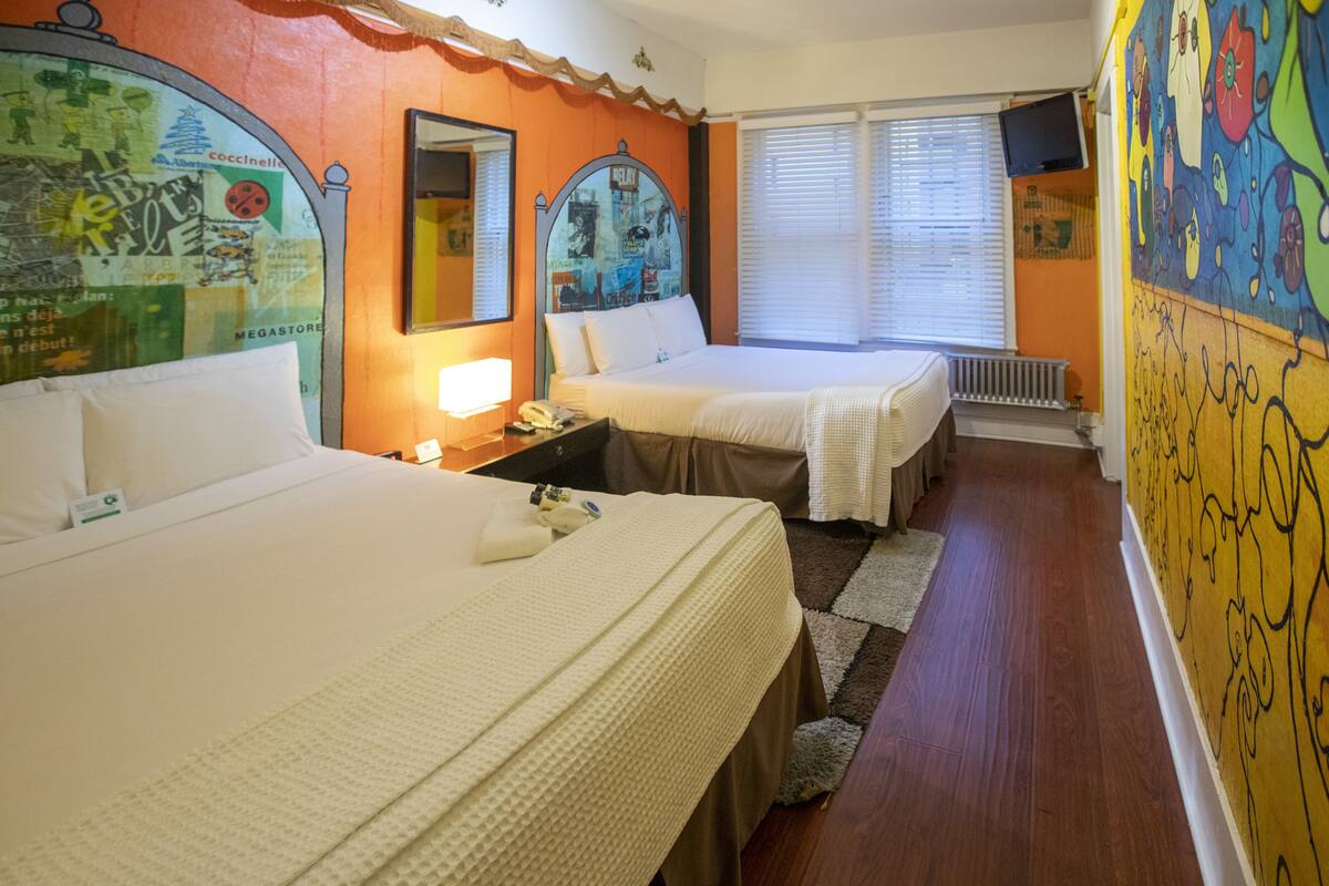 A hotel room with two beds, a large mirror, a window with blinds, and a colorful wall mural.