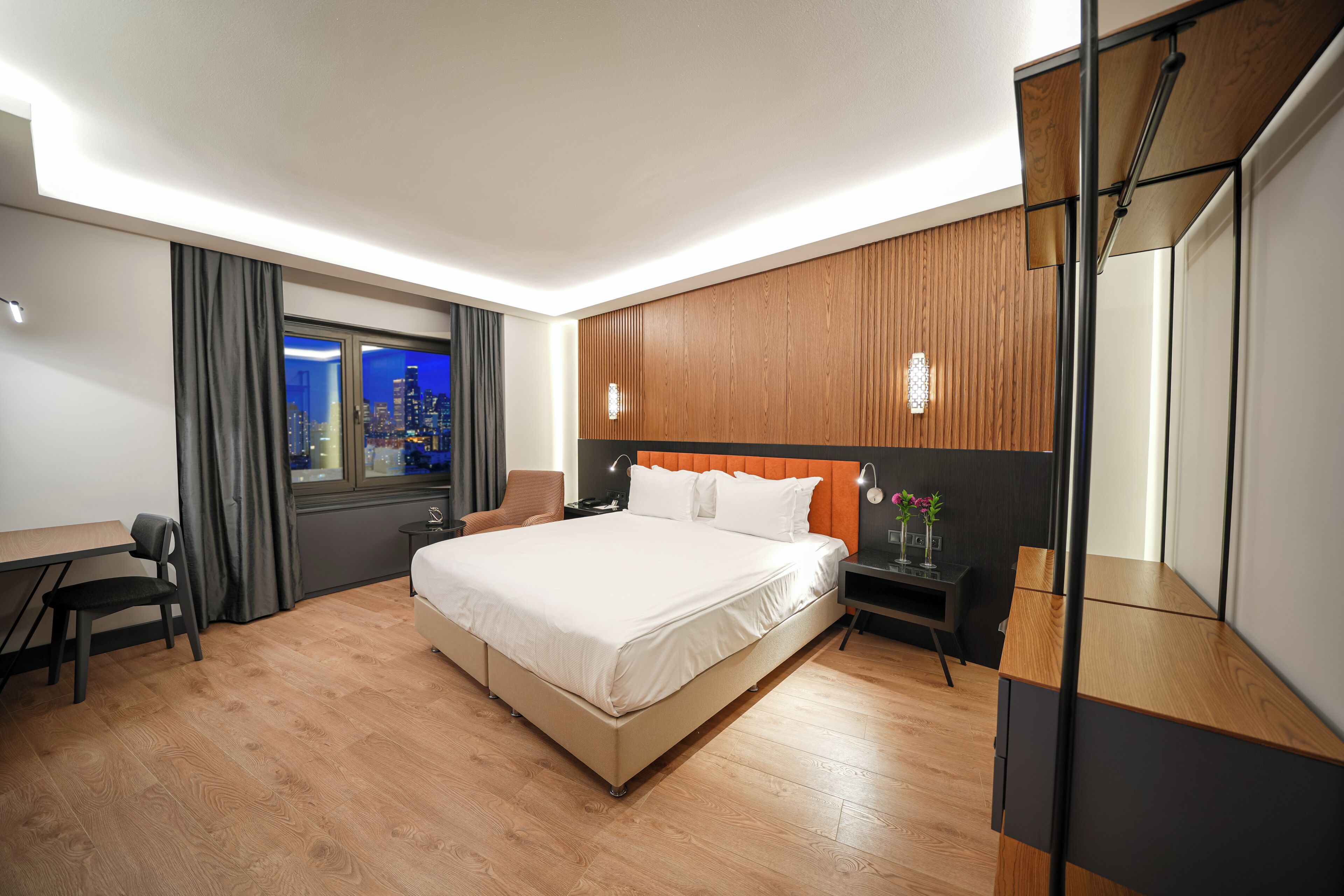 Una camera d'albergo moderna con un letto grande, una scrivania, una sedia, un comodino e vista sulla città attraverso una finestra.