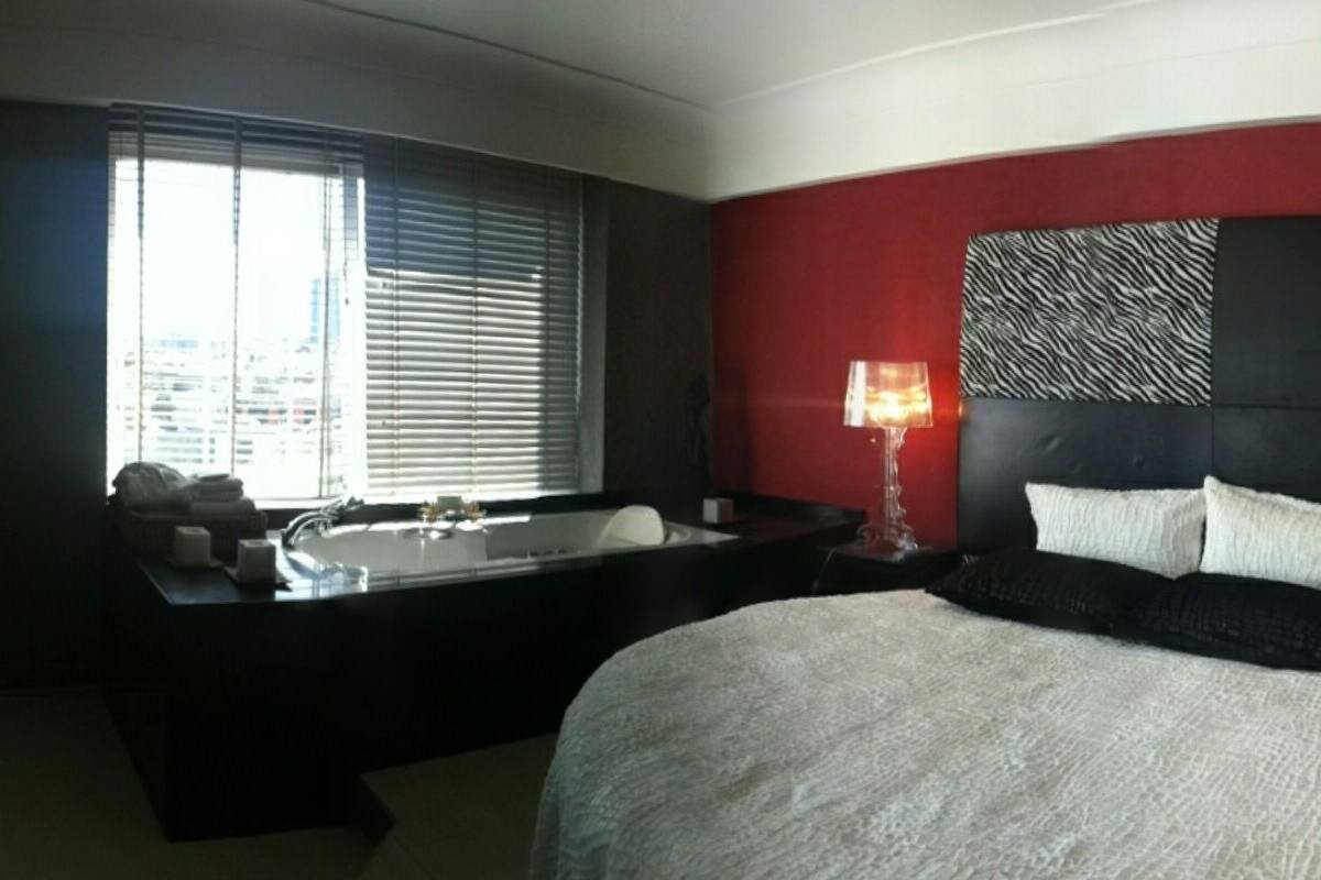 A hotel room with a large bed, a separate bathtub, a red accent wall, and a city view through the window.