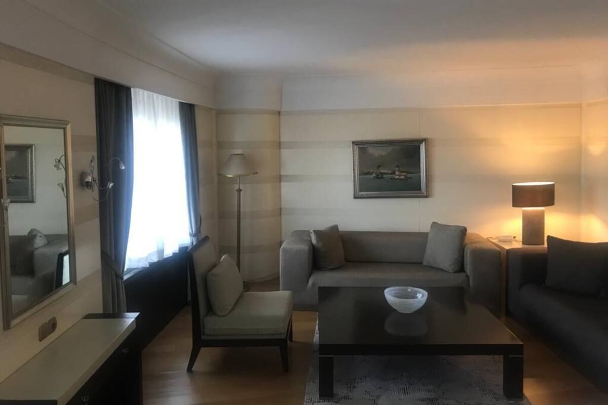 A modern hotel room with a sofa, armchair, and a coffee table. There is a large mirror, a lamp, and a painting on the wall.