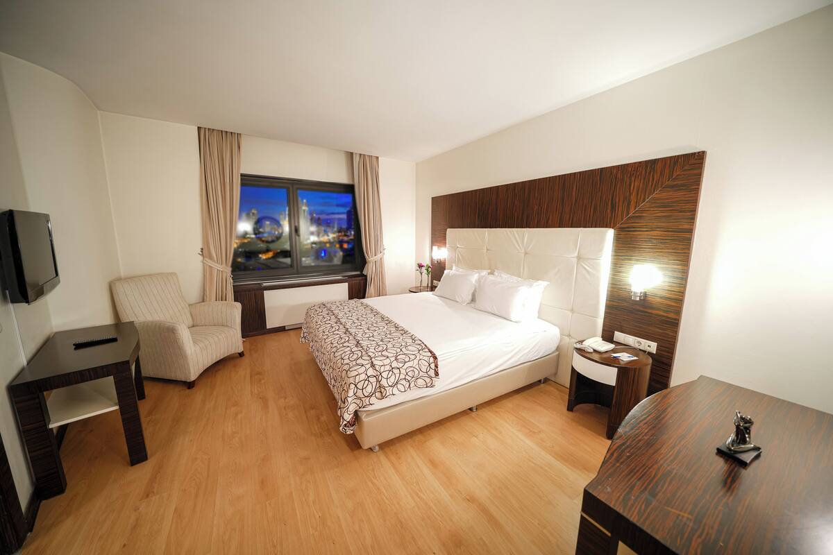 A hotel room with a bed, a chair, a desk, a TV, and a window with a city view.