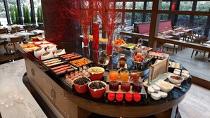 A buffet with various food items and beverages, a large pillar with red decorations, and a view of the outdoors through large windows. - Surmeli Istanbul Hotel (Istanbul)