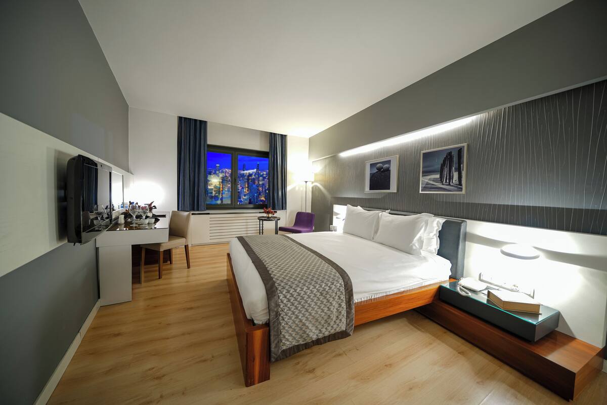 A modern hotel room with a bed, bedside table, lamp, and a desk with a chair.