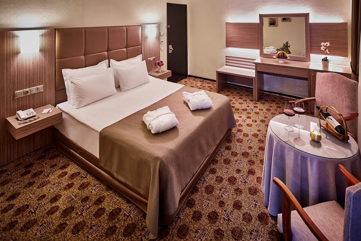 A hotel room with a bed, bedside table, a desk with a chair, a round table with a chair, and a mirror.