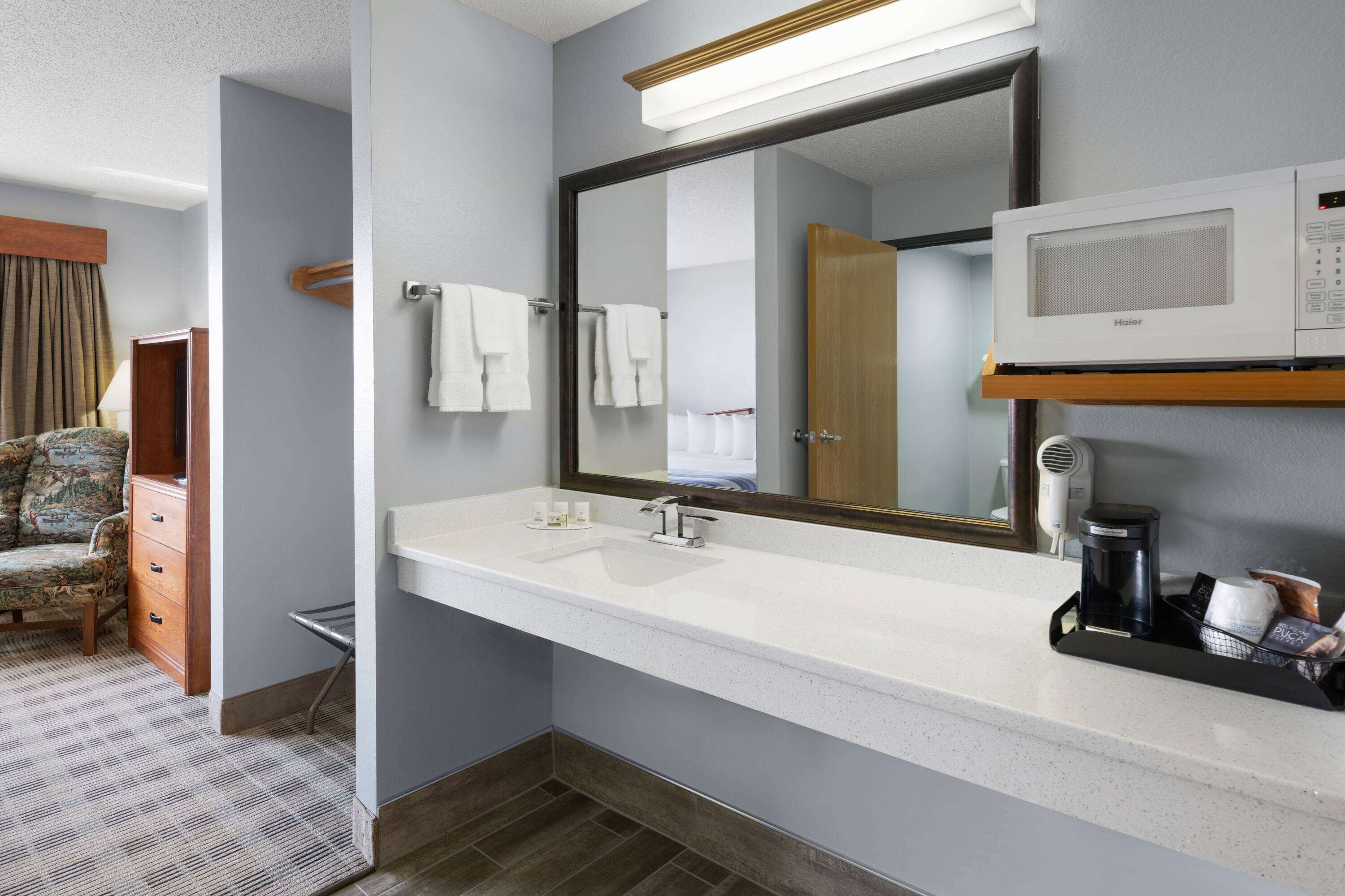 A hotel bathroom with a large mirror, a white sink, a towel rack, a microwave, and a coffee maker.