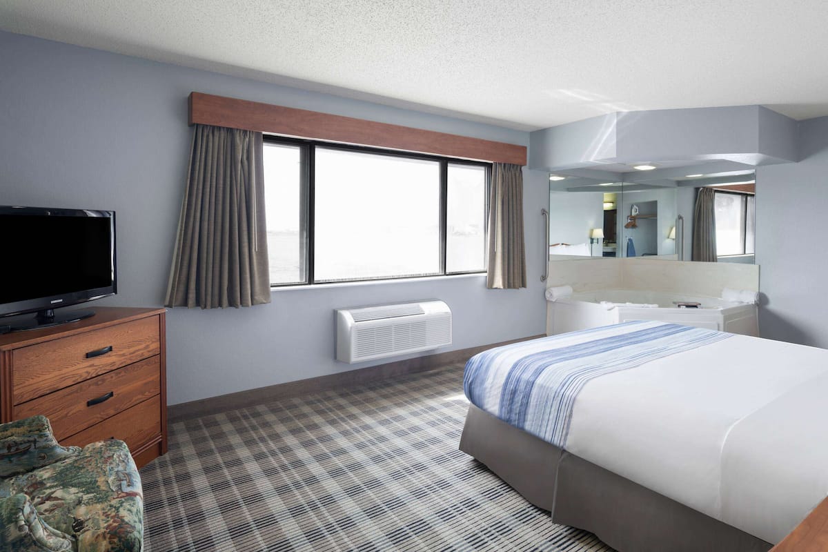 A hotel room with a bed, a television on a wooden dresser, a window with curtains, a mirror, and a bathroom with a bathtub and sink.