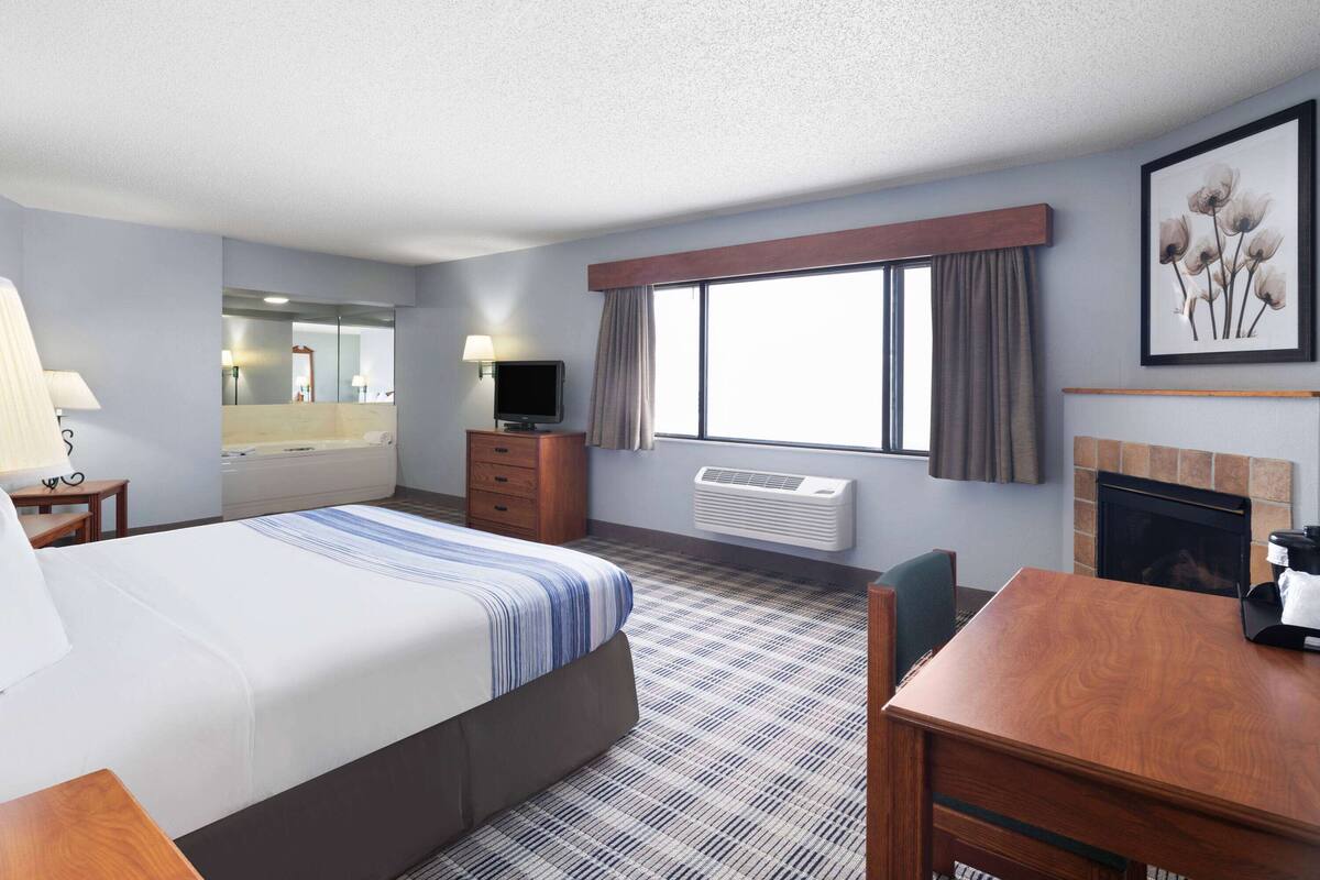 A hotel room with a bed, a desk, a television, a mirror, a fireplace, and a large window with curtains.