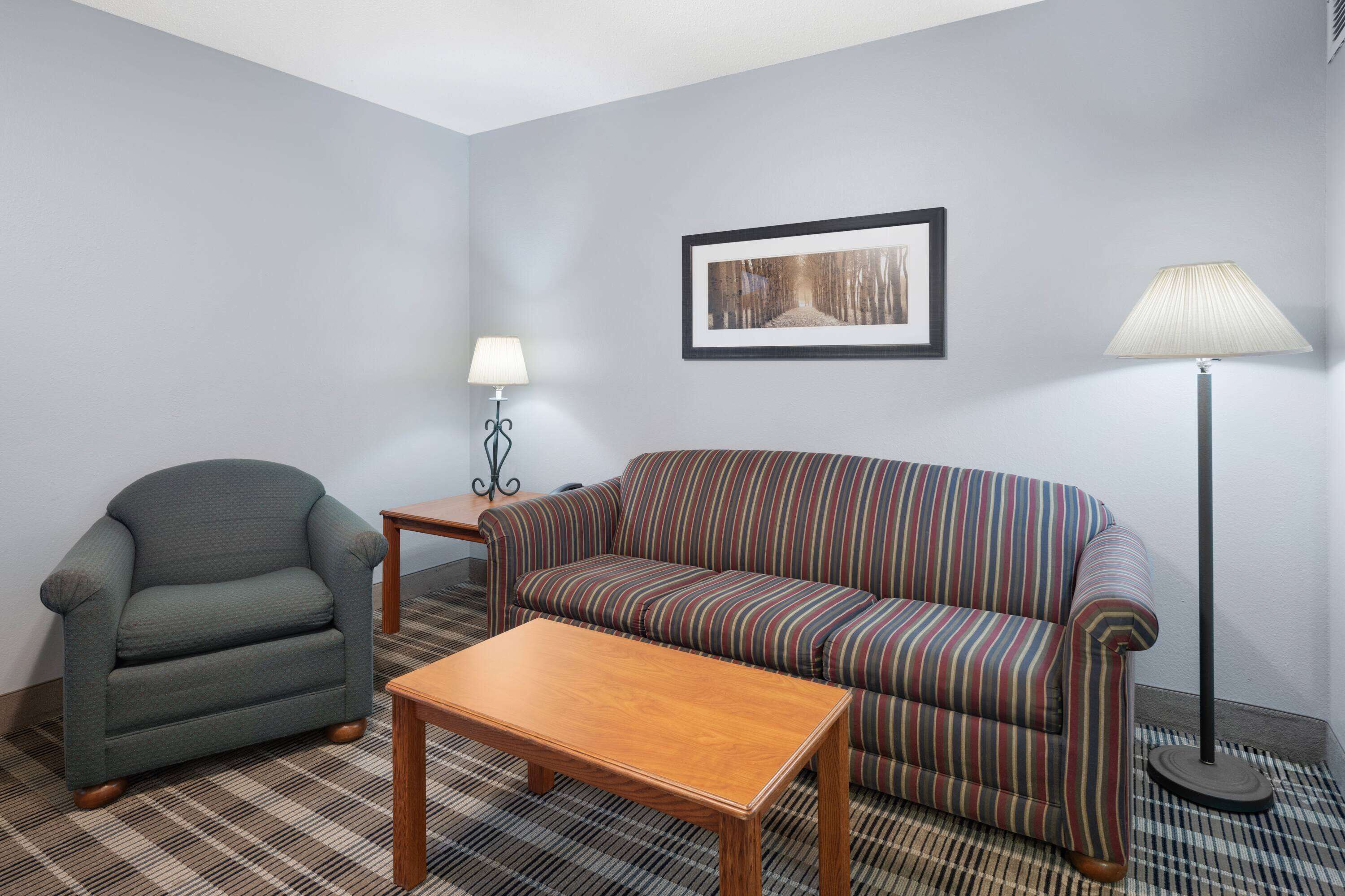 A room with a striped sofa, a green armchair, a wooden coffee table, a floor lamp, and a framed picture on the wall.
