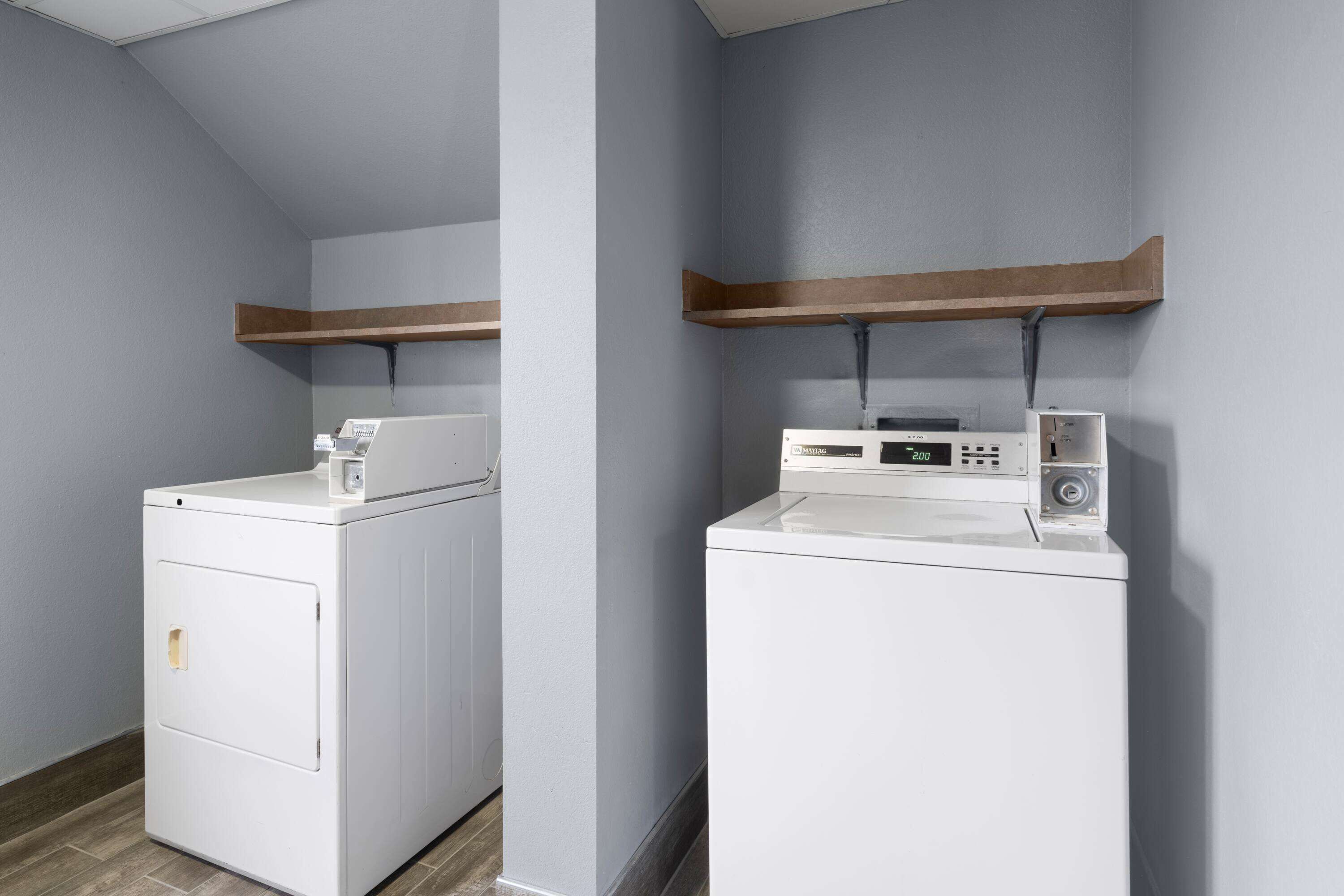 A laundry room with two white washing machines, wooden shelves above them, and a grey wall.