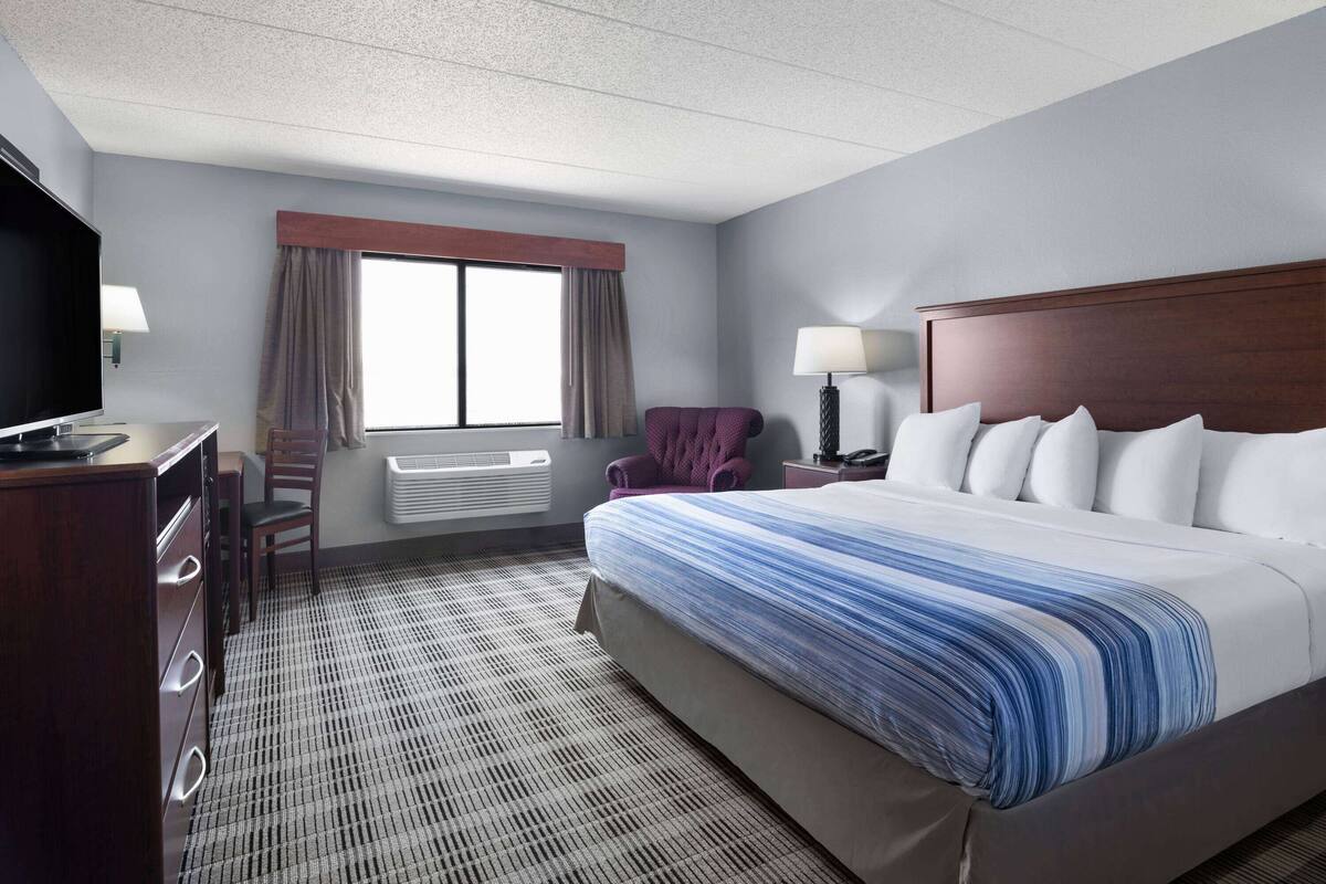 A hotel room with a large bed, a desk with a television, a chair, a window with curtains, and a nightstand with a lamp.
