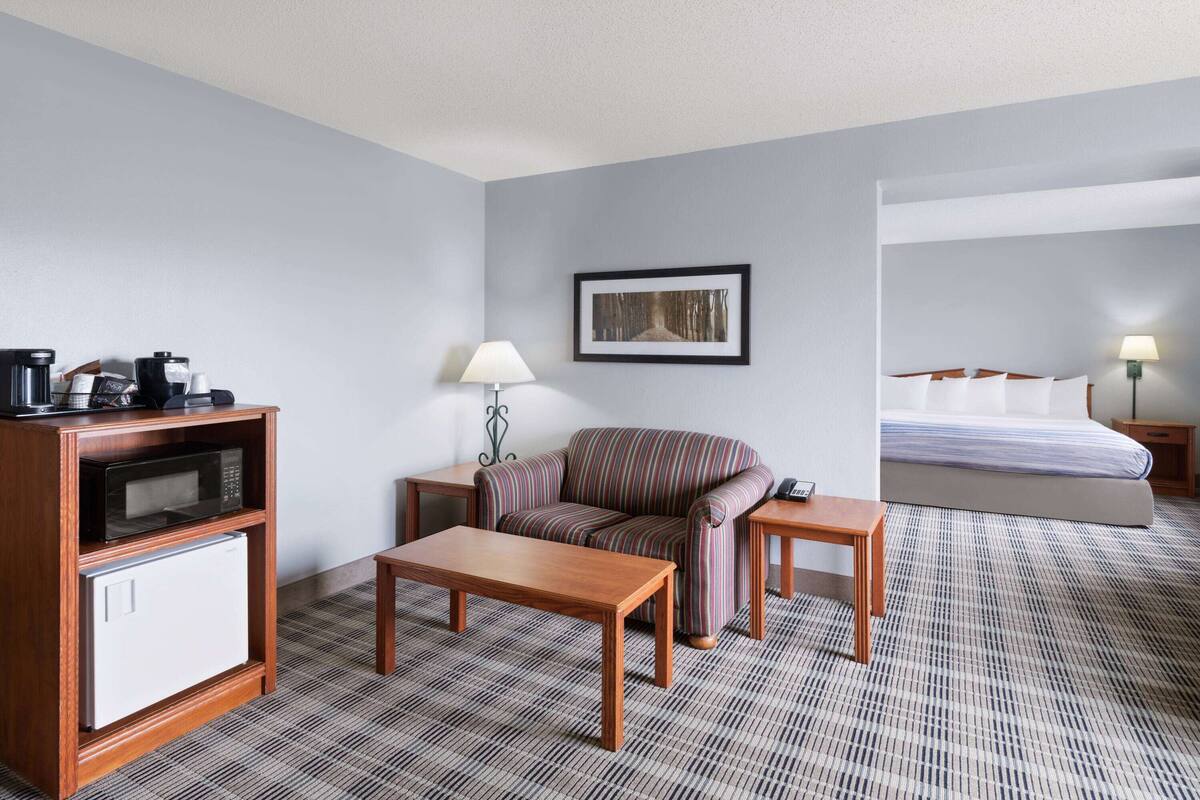 A hotel room with a sofa, a small table, a coffee machine, a microwave, a lamp, and a bed with a pillow.