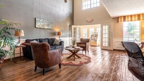 A spacious living room with a high ceiling, a ceiling fan, a sofa, armchairs, a coffee table, and a large window providing natural light.