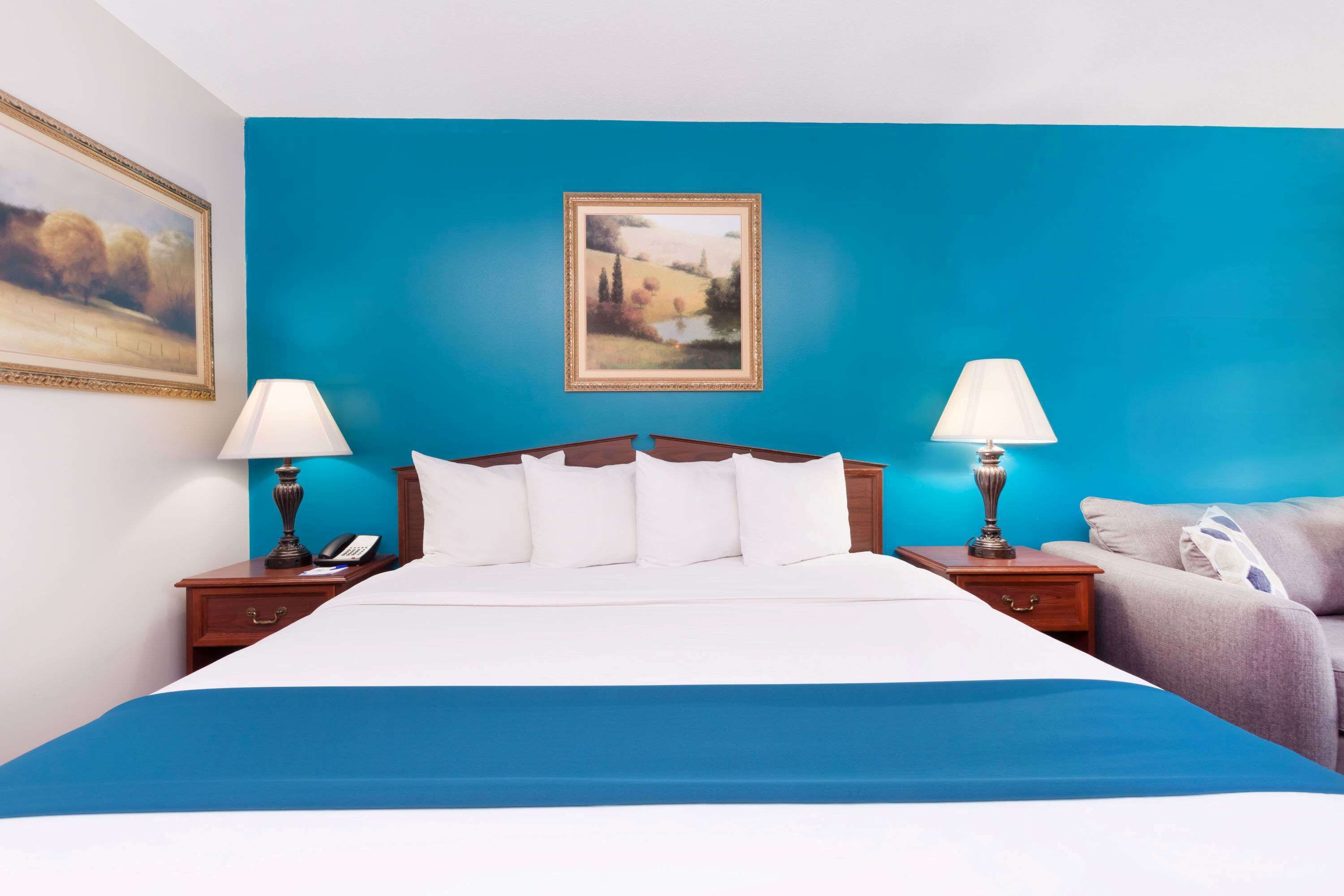 A hotel room with a large bed, two bedside tables with lamps, a painting on the wall, and a view of the outdoors.