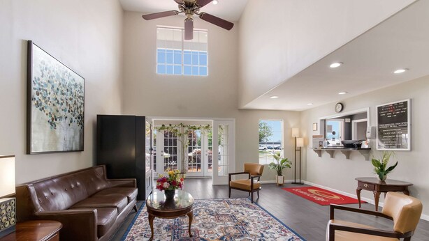 A spacious lobby with a high ceiling, a large window, a ceiling fan, a brown leather sofa, a glass coffee table with flowers, a patterned rug, and a red carpet. - Econo Lodge Greenville near University (Greenville)