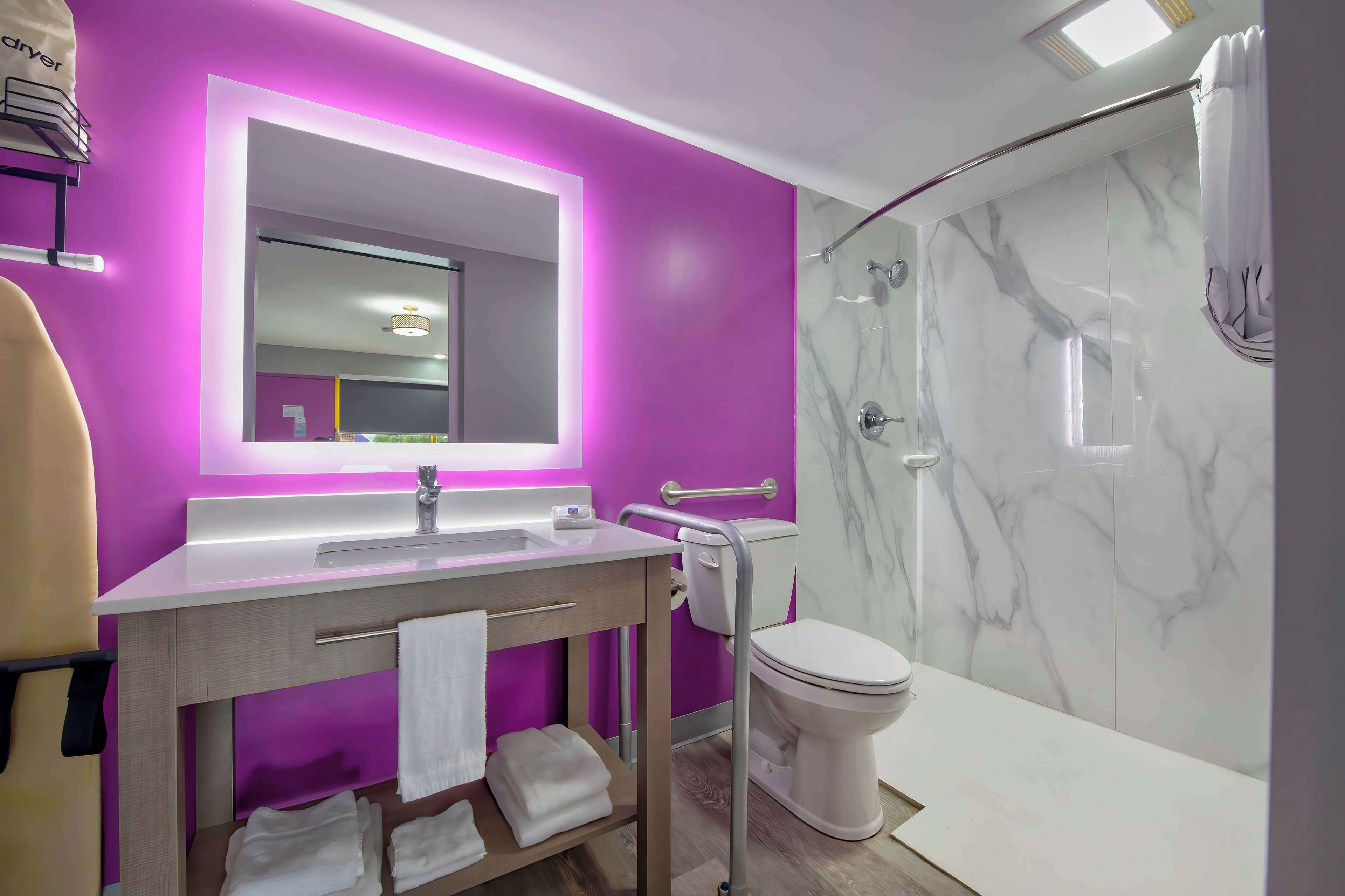 Un baño moderno con pared morada, un espejo grande, un lavabo blanco y una zona de ducha con azulejos de mármol.