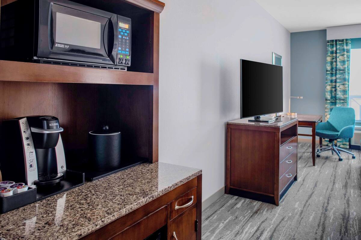 A hotel room with a kitchenette, a flat-screen TV, a desk, and a chair.