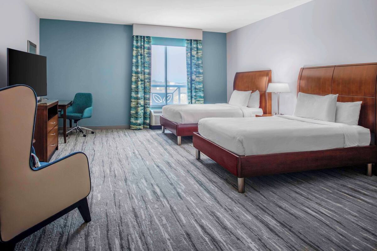 A hotel room with two beds, a desk with a chair, a TV, and a large window with curtains.