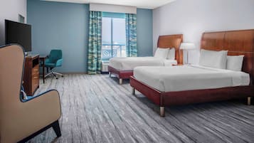 Quarto de hotel com duas camas, uma mesa de trabalho com cadeira, televisão e uma janela grande com cortinas.