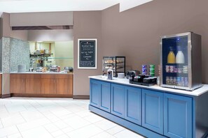 Un área de cocina moderna con barra americana, un refrigerador con bebidas y un menú en pizarra que anuncia los horarios del desayuno y un desayuno tipo buffet completo.