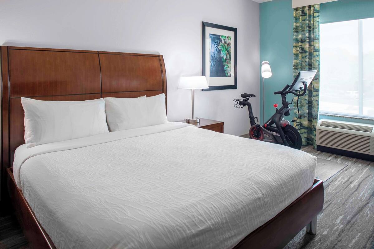 A hotel room with a large bed, a nightstand, a lamp, a bicycle, and an air conditioning unit.