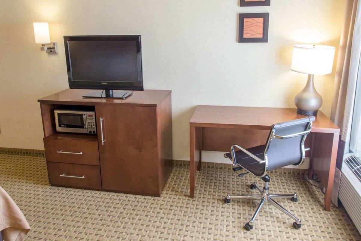 A hotel room with a flat-screen TV on a wooden cabinet, a desk with a chair, a lamp, and two framed pictures on the wall.