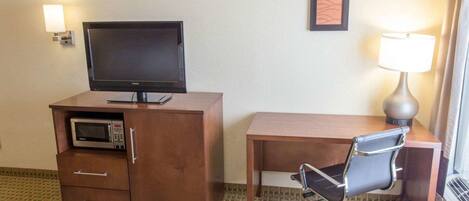 A hotel room with a flat-screen TV on a wooden cabinet, a desk with a chair, a lamp, and two framed pictures on the wall.