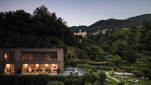 A modern building with a pool and lounge area, surrounded by lush greenery and hills in the background.