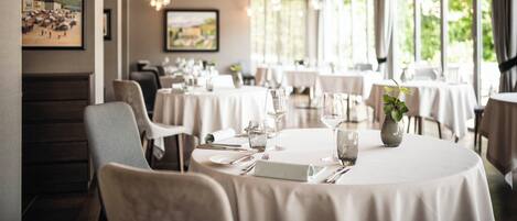 A restaurant with round tables, white tablecloths, and hanging light fixtures.