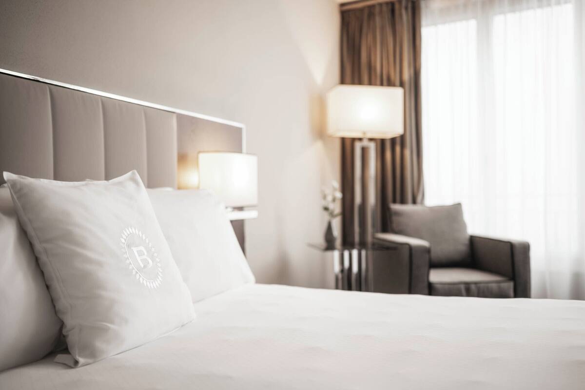 A hotel room with a bed, a chair, a lamp, and a window with curtains.