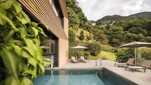 A pool area with lounge chairs and umbrellas, surrounded by greenery and hills.