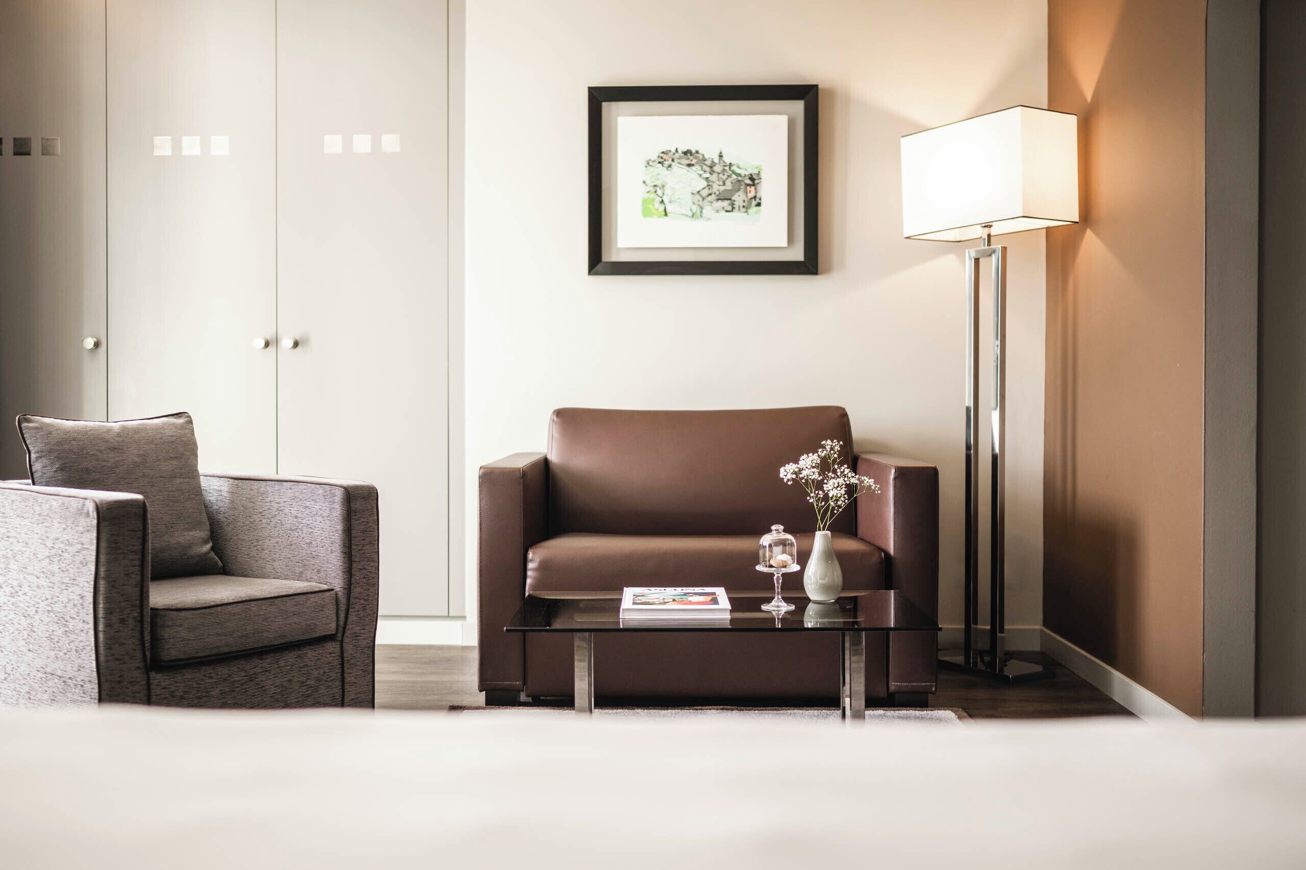 Une chambre d’hôtel moderne avec un canapé en cuir marron, une table basse en verre sur laquelle reposent un magazine et un vase, une lampe sur pied et un tableau encadré au mur.