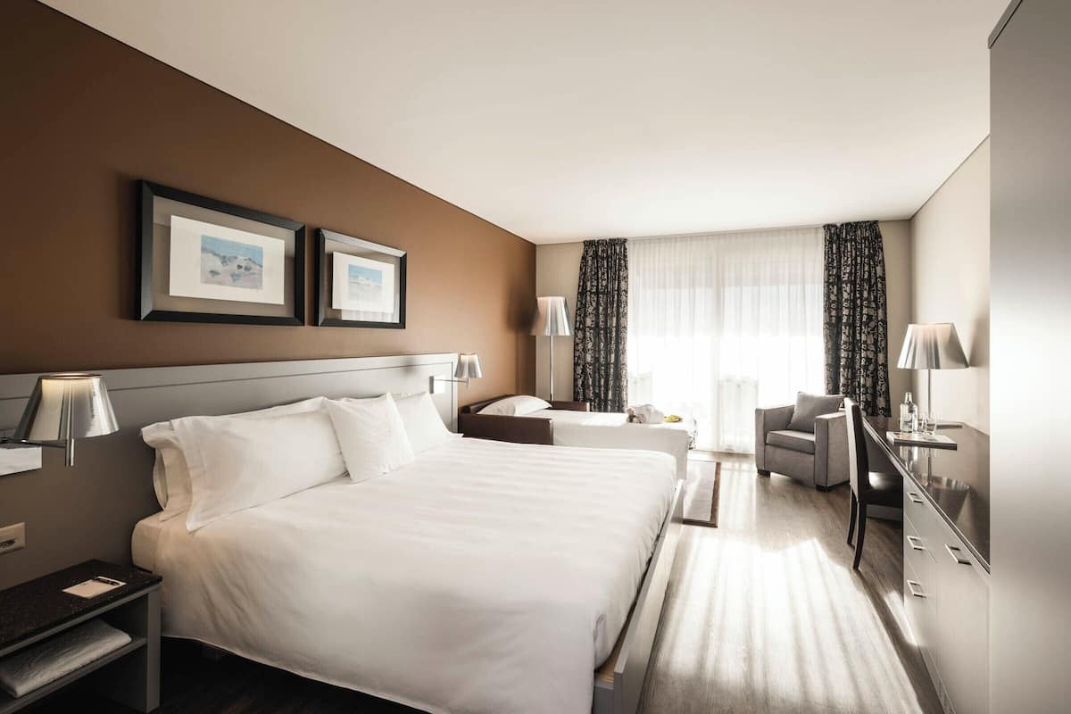 A hotel room with a large bed, bedside tables, a desk, and a chair. There are two framed pictures on the wall, a window with curtains, and a lamp on the desk.