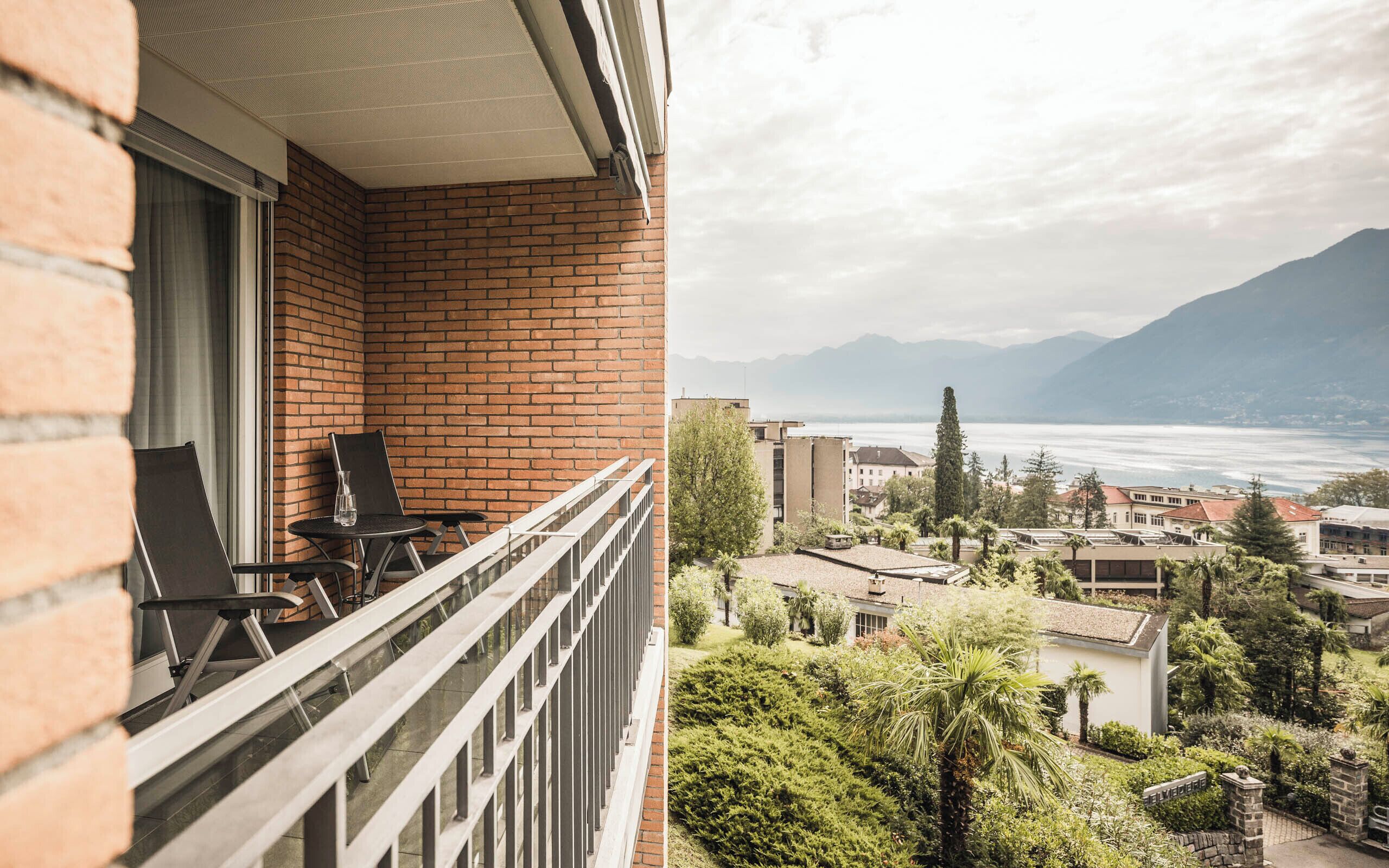 A balcony with a brick wall, two chairs, and a table. A view of a lake and mountains is visible through the window.